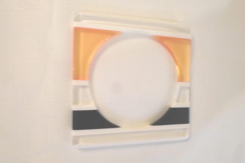 May include: A square white plastic light switch cover plate featuring a central circular cutout. The design includes a horizontal orange rectangular accent at the top and a horizontal black rectangular accent at the bottom of the plate.