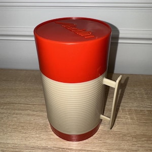 May include: A vintage Aladdin thermos with a red top and base, and a cream-colored, ribbed body. The handle is cream-colored. The word "Aladdin" is embossed on the lid. This retro thermos is a classic for picnics and outdoor adventures.