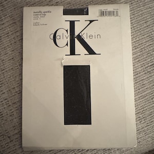 May include: A package of Calvin Klein metallic sparkle control top pantyhose in black and silver. The package is white with the Calvin Klein logo in black and the letter "K" in a large, bold font. The package also includes the style number, size, and color.
