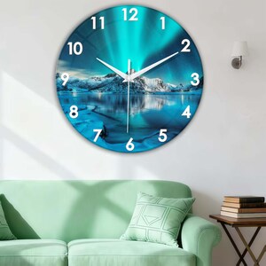 Könnte beinhalten: Eine runde Wanduhr mit einem Landschaftsdesign. Das Zifferblatt zeigt eine blau-weiße Szene des Nordlichts mit einer Bergkette und einem Gewässer. Weiße Zeiger zeigen die Uhrzeit an. Die Uhr ist an einer weißen Wand befestigt.