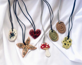 May include: A selection of handmade ceramic pendant necklaces. The pendants showcase diverse shapes, such as a starburst, heart, mushroom, and spiral designs. Each pendant is suspended on a black or grey cord, set against a white fabric backdrop.