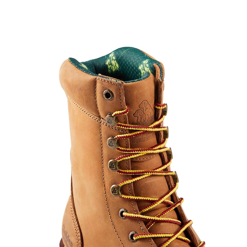 May include: Close-up of a tan work boot with red and yellow laces. The boot has a padded collar with a green and yellow patterned lining. The boot features metal eyelets and a logo on the side.