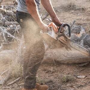 May include: An orange and white STIHL chainsaw cutting through a log. The image shows a person wearing brown work boots and dark pants. Sawdust fills the air as the chainsaw cuts through the wood.