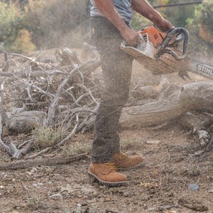 May include: An orange and white STIHL chainsaw cutting through a log. The operator is wearing work boots and dark pants. Sawdust and debris are visible in the air, indicating the cutting process. The scene takes place outdoors.
