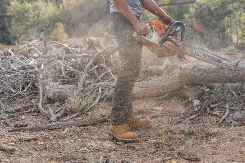 May include: An orange and white STIHL chainsaw cutting through a log. The operator is wearing work boots and dark pants. Sawdust and debris are visible in the air, indicating the cutting process. The scene takes place outdoors.