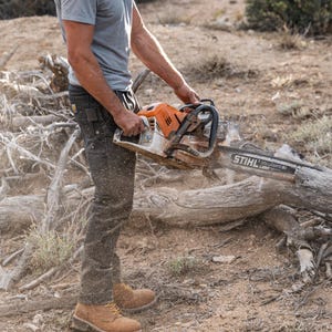 May include: An orange and silver STIHL chainsaw being used outdoors. The chainsaw is held by a person wearing a gray t-shirt, dark pants, and work boots. The person is wearing a helmet with ear protection. The ground is covered in dirt and wood debris.