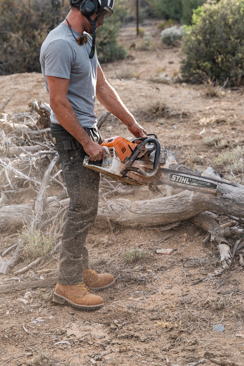 May include: An orange and silver STIHL chainsaw being used outdoors. The chainsaw is held by a person wearing a gray t-shirt, dark pants, and work boots. The person is wearing a helmet with ear protection. The ground is covered in dirt and wood debris.