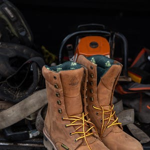 May include: A pair of brown leather work boots with yellow and brown laces. The boots are waterproof and have a rugged sole. The boots are sitting in the back of a truck with other work equipment, including a chainsaw.
