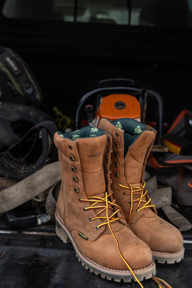 May include: A pair of brown leather work boots with yellow and brown laces. The boots are waterproof and have a rugged sole. The boots are sitting in the back of a truck with other work equipment, including a chainsaw.