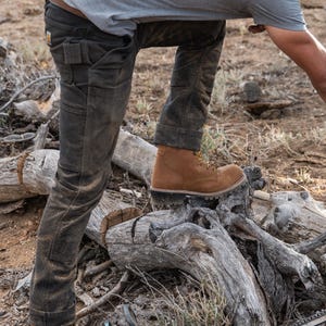 May include: A person wearing brown work boots with yellow laces and dark work pants stands on a pile of logs. An orange chainsaw with the text "STIHL" is in the foreground. The person is wearing a gray t-shirt.