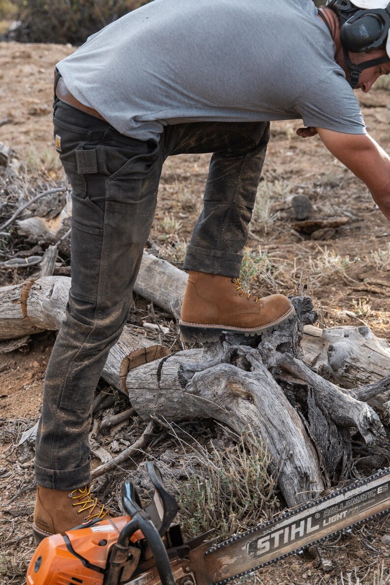 May include: A person wearing brown work boots with yellow laces and dark work pants stands on a pile of logs. An orange chainsaw with the text "STIHL" is in the foreground. The person is wearing a gray t-shirt.