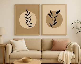 May include: Two framed botanical art prints. Each print features a black leaf design on a beige circle, set against a tan background. The prints are displayed above a beige sofa with pillows and a blanket, creating a warm, neutral-toned living room.