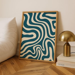 May include: A framed art print featuring a teal and cream wavy pattern. The artwork is displayed on a wooden floor, leaning against a wall. A gold-colored lamp and books are visible.