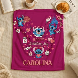 A pink blanket with a Disney character design and the text "Happy Valentine's day" printed on it, along with the name "Carolina" written below. The blanket is placed on a bed, with a newspaper, a croissant, and a mug visible in the background.
