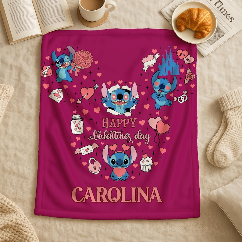 A pink blanket with a Disney character design and the text "Happy Valentine's day" printed on it, along with the name "Carolina" written below. The blanket is placed on a bed, with a newspaper, a croissant, and a mug visible in the background.