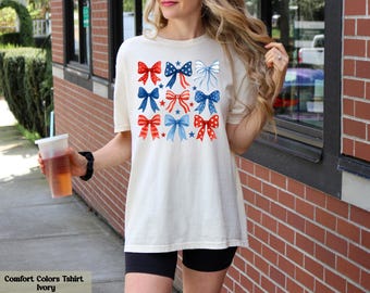 May include: Ivory Comfort Colors t-shirt featuring a pattern of red, white, and blue bows. The bows are in various styles and are scattered across the front of the shirt. The shirt is paired with black shorts.