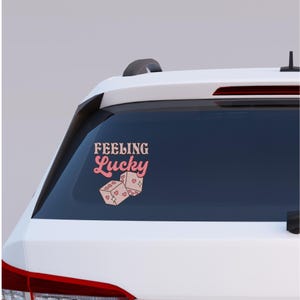 May include: A pink and beige sticker on a car window. The sticker features the text "Feeling Lucky" in a retro font above two dice with heart symbols. The dice are beige with pink hearts and dots.