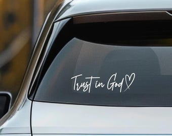 May include: White vinyl decal applied to a car window. The decal features the handwritten phrase "Trust in God" with a heart symbol. The decal is a decorative item for vehicles, promoting a message of faith and belief.