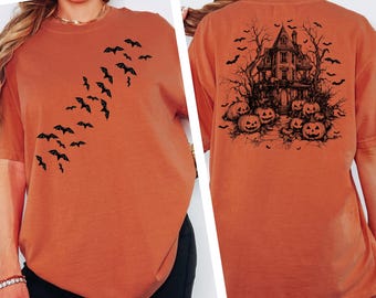 May include: Orange Comfort Colors T-shirt with black bat silhouettes on the front and a haunted house with pumpkins on the back. The shirt is a relaxed fit, perfect for Halloween.