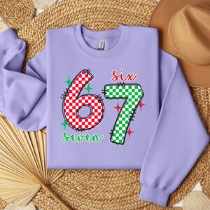 a purple sweatshirt with the number "67" and the text "seven" printed on it, along with a brown hat and a straw hat placed on a woven mat.
