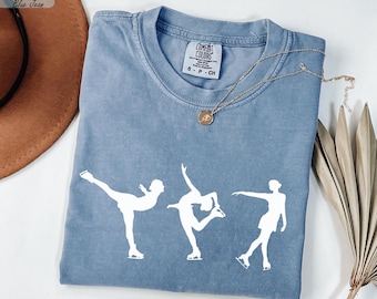 May include: A blue jean Comfort Colors t-shirt featuring three white ice skating figures in motion. The shirt has a crew neck and short sleeves. A gold necklace with a pendant is draped across the shirt.