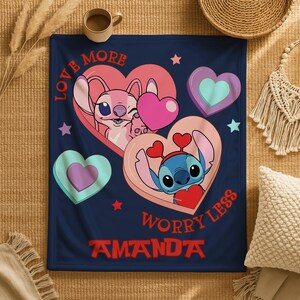A blanket with a design featuring two cartoon characters, one pink and one blue, surrounded by hearts and stars. The blanket has the text "LOVE MORE WORRY LESS" and "AMANDA" printed on it.