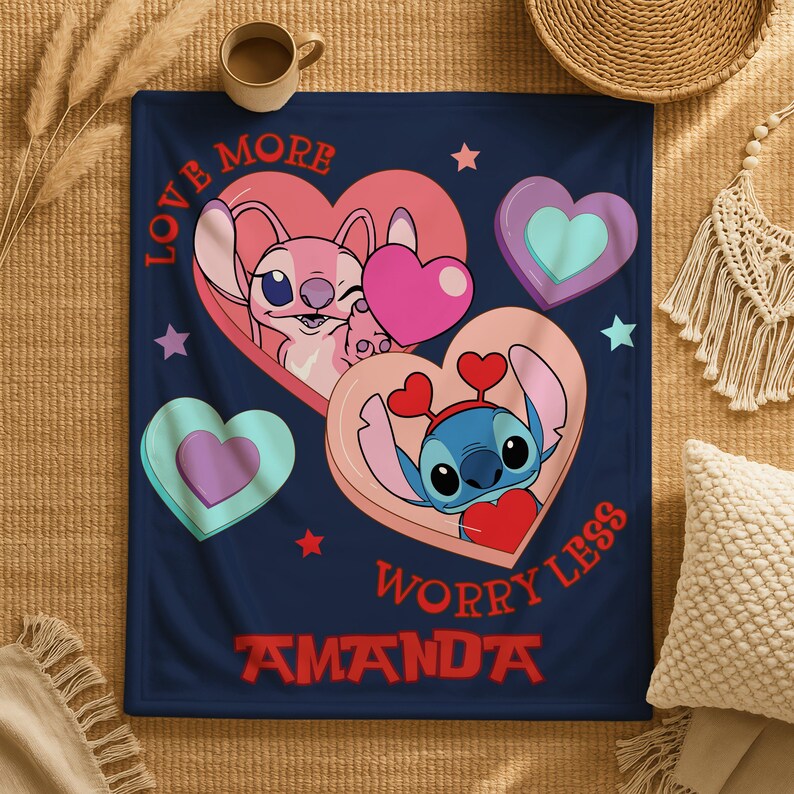 A blanket with a design featuring two cartoon characters, one pink and one blue, surrounded by hearts and stars. The blanket has the text "LOVE MORE WORRY LESS" and "AMANDA" printed on it.