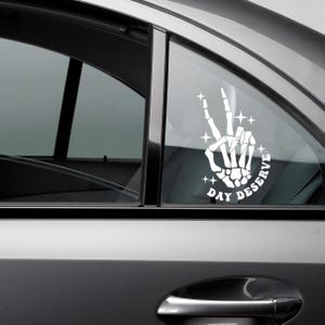 May include: White vinyl car decal featuring a skeleton hand making a peace sign. The text "DAY DESERVE" is arched around the base of the hand, with star accents. The decal is applied to a car window.