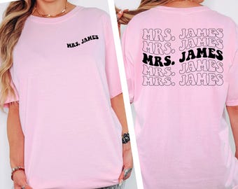 May include: A light pink t-shirt with the text "MRS. JAMES" on the front and back. The back of the shirt has the text "MRS. JAMES" repeated in a stacked design, with one instance in black. The shirt is a comfort colors brand.