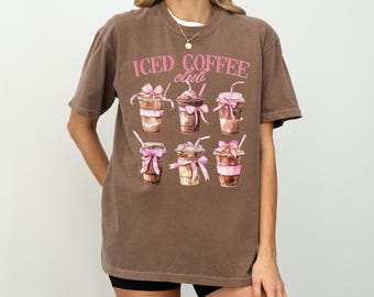 May include: Brown t-shirt with the text "ICED COFFEE club" in pink. The shirt features a watercolor design of six iced coffee drinks with pink bows and straws. The words "Comfort Colors Espresso" are at the bottom of the shirt.