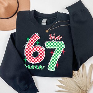 A black sweatshirt with a large number "67" printed on it, along with a green and red checkered pattern. The sweatshirt is placed on a white surface, and there is a brown hat and a fan nearby.