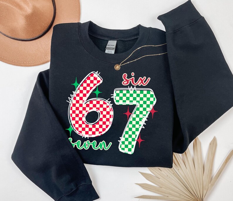 A black sweatshirt with a large number "67" printed on it, along with a green and red checkered pattern. The sweatshirt is placed on a white surface, and there is a brown hat and a fan nearby.