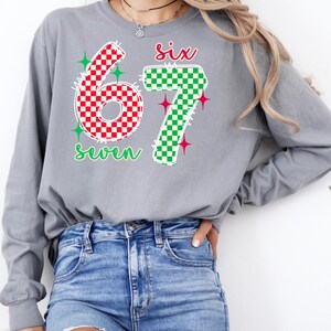 A woman wearing a gray long-sleeved shirt with a graphic design featuring the numbers '67' and '7' in red and green checkered patterns, along with the text six seven'. She is also wearing distressed blue jeans.