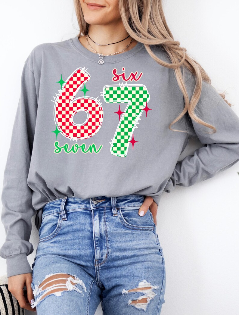 A woman wearing a gray long-sleeved shirt with a graphic design featuring the numbers '67' and '7' in red and green checkered patterns, along with the text six seven'. She is also wearing distressed blue jeans.
