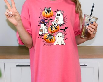 May include: A pink t-shirt featuring a Halloween-themed graphic. The design includes cartoon ghosts, pumpkins with bows, spiderwebs, and bats. The text "Cc Watermelon" is visible at the bottom left.