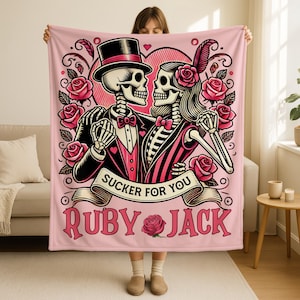 May include: Pink blanket featuring a skeleton couple in formal attire, surrounded by roses and a heart. The blanket has the text "Sucker for You" and "Ruby & Jack". The skeletons are in a romantic embrace, with the male skeleton wearing a top hat.