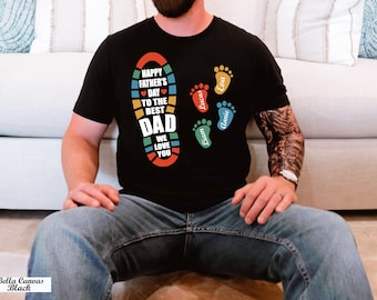 May include: Black t-shirt with a colorful graphic that reads "Happy Father's Day to the Best Dad, We Love You." The design includes a footprint and the names Lucas, Eva, Annie, and Liam in colorful footprints.