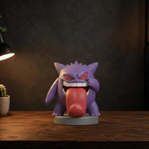 Può includere: Una statuetta viola di Gengar posizionata su una base circolare grigia. Il personaggio presenta orecchie a punta, occhi rossi stretti, una bocca larga con denti bianchi e una lunga e prominente lingua rosa che fuoriesce.
