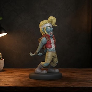 May include: A figurine of a blue-skinned zombie character with blonde hair tied in a ponytail and a white headband. The figure wears a red vest over a white shirt, tan pants with distressed details, and grey boots, standing on a circular black base.