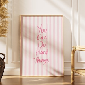 May include: A framed print with a pink and white striped background. The text "You Can Do Hard Things" is written in pink script. The print is leaning against a white wall, next to a vase with dried plants.