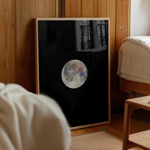 May include: A framed print showing a colourful depiction of the moon against a black backdrop. The moon displays various colours, including blue, red, and yellow, set within a wooden frame. The artwork is displayed in a room with wooden panelling and a bed.