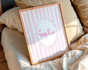 May include: A framed print featuring the name "Sophie" in pink script within a decorative oval. The background has vertical pink and white stripes. The frame is made of light-colored wood. The print is displayed on a bed with pillows and a comforter.