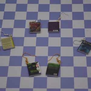 Puede incluir: Pendientes en miniatura con forma de libro, con ganchos plateados y dorados. Los pendientes presentan portadas de libros como "Harry Potter" y "Wicked", sobre un fondo a cuadros azul y blanco.