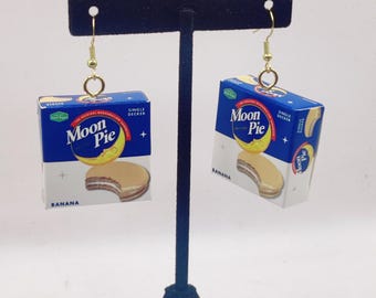 May include: Novelty earrings shaped like miniature Moon Pie boxes. The boxes are blue and white, displaying the Moon Pie logo and a banana-flavored treat image. The earrings are suspended from gold-coloured hooks.