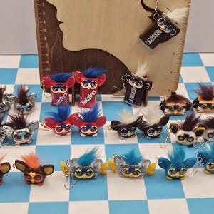 May include: A collection of handmade Furby-inspired earrings in various colors and styles. Some earrings are silver, red, brown, and blue, with fluffy hair and large eyes. Some earrings are designed to look like Hershey's chocolate bars and Twizzlers.