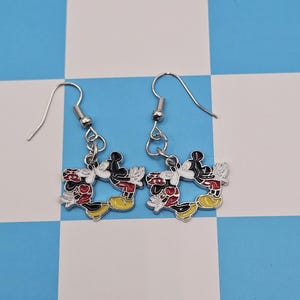 May include: A pair of silver-toned dangle earrings. Each earring features a colorful enamel design of Mickey Mouse in a classic pose. The earrings have red, black, yellow, and white accents, with a fish hook closure.