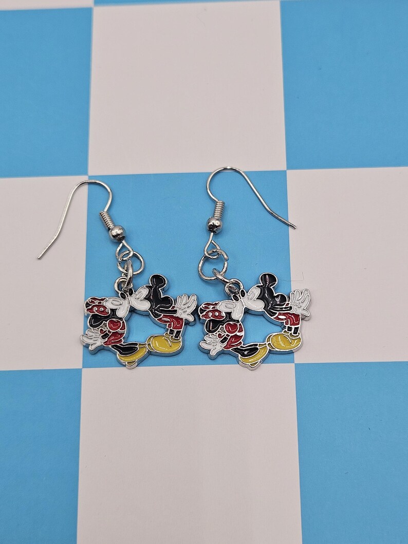 May include: A pair of silver-toned dangle earrings. Each earring features a colorful enamel design of Mickey Mouse in a classic pose. The earrings have red, black, yellow, and white accents, with a fish hook closure.