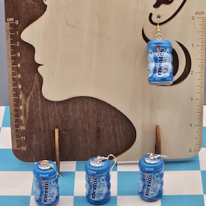 Puede incluir: Pendientes de chicle Mentos azules en miniatura con ganchos dorados. Los pendientes son réplicas de los envases de chicle, con el logo "mentos PURE FRESH". Se exhiben sobre un recorte de madera con forma de rostro y una regla en cm y pulgadas.