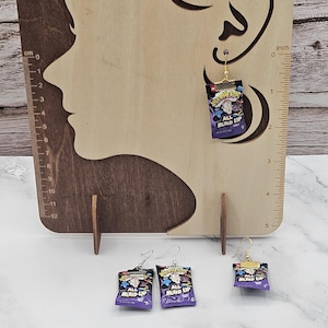 May include: Three dangle earrings featuring miniature purple Airheads All Mixed Up candy wrappers. Each earring has a gold-tone metal top and a gold-tone hook. The earrings are displayed on a wooden board with a ruler showing a height of 1.5 inches.