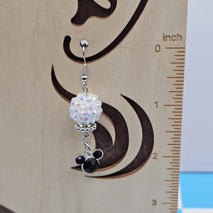 May include: A single silver earring with a white, iridescent, beaded ball and a black Mickey Mouse charm. The earring is hanging in front of a wooden background with a ruler showing inches.